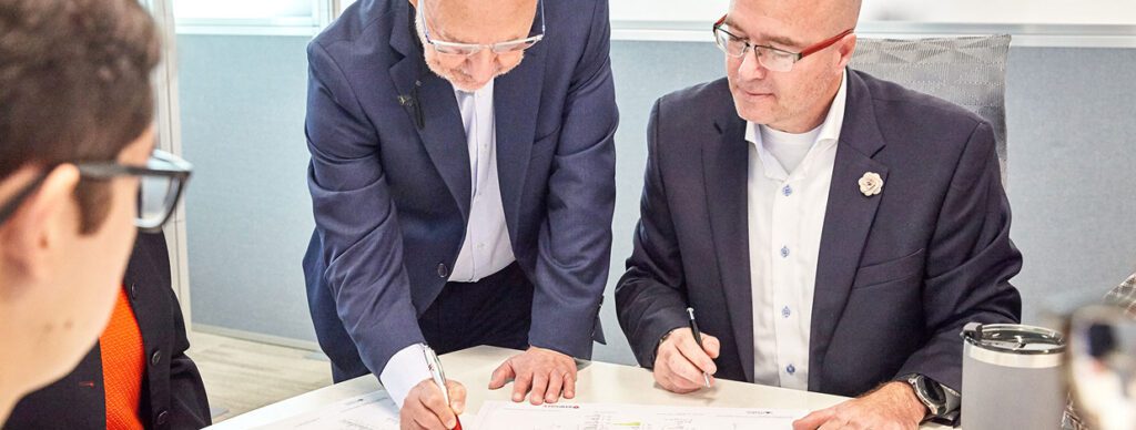 Two business professionals in formal attire reviewing and signing technical documents during a corporate meeting