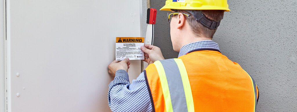 Electrician wearing safety gear placing an arc flash warning label on an electrical panel to ensure workplace safety compliance