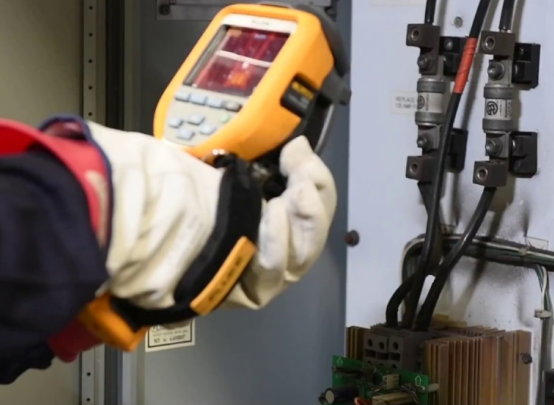 echnician wearing protective gloves using a handheld thermal imaging camera to inspect components inside an electrical panel.
