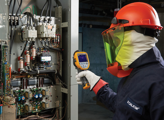 Electrical technician in full arc flash protective gear using a thermal imaging camera to inspect components inside an industrial control panel.